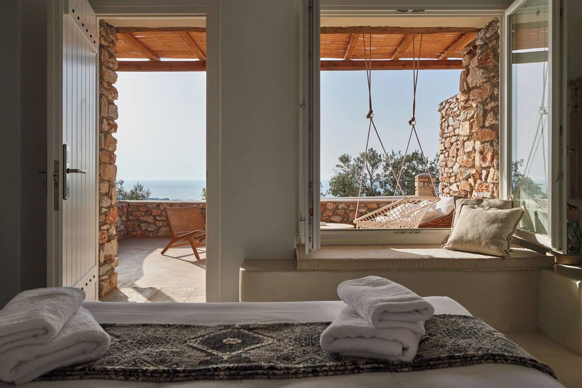 A room with a bed, a view of the sea, a stone wall, and a wooden ceiling.