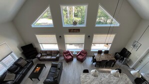 A spacious living room with a high ceiling, large windows, a dining area, and a fireplace. - Kinabik Cabin / Half block from beach, quiet cottage district (Sylvan Lake)