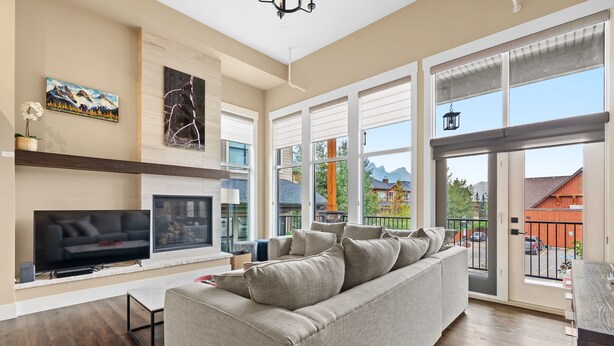 A spacious living room with a large gray sofa, a flat-screen TV, a fireplace, and large windows offering a view of the outdoors.