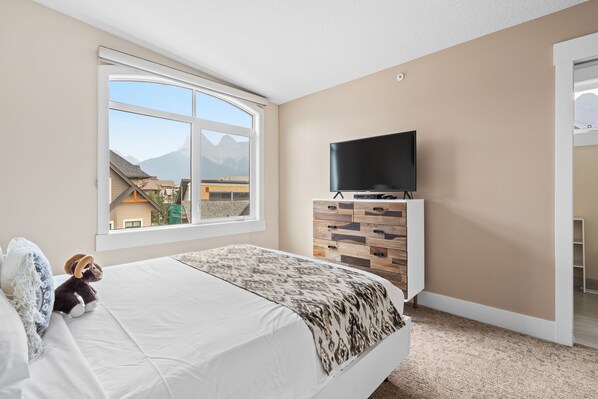 A bedroom with a large window, a bed with a patterned blanket, a television on a wooden cabinet, and a view of buildings and mountains. - Mountain Views! Art! Luxurious 5Brs/4.5Ba Townhome, Hot Tub, Canmore for 12 (Canmore)