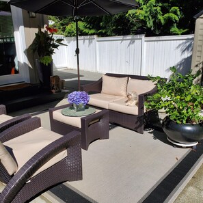 An outdoor patio area with wicker furniture, a cushioned sofa, a round table with a vase of flowers, and a large potted plant.