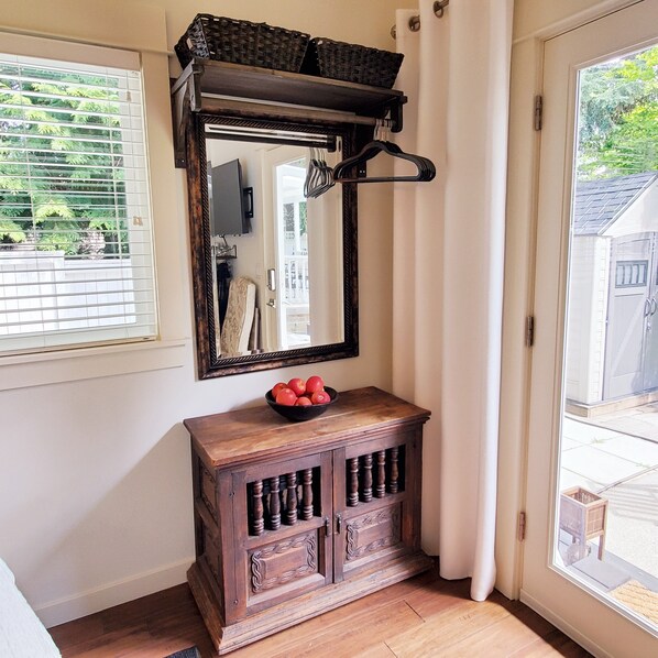 A wooden cabinet with a mirror above it, a bowl of apples on the cabinet, and a window with blinds.