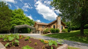 A log cabin with a stone chimney, surrounded by a well-maintained garden and a covered patio area. - Log home on 50 acres. Private, convenient, beautiful. A little bit of paradise. (Knoxville)