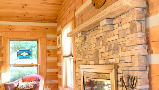 A log cabin interior with a stone fireplace, a mounted deer head, and a cozy seating area with a leather chair and a small table. - Log home on 50 acres. Private, convenient, beautiful. A little bit of paradise. (Knoxville)