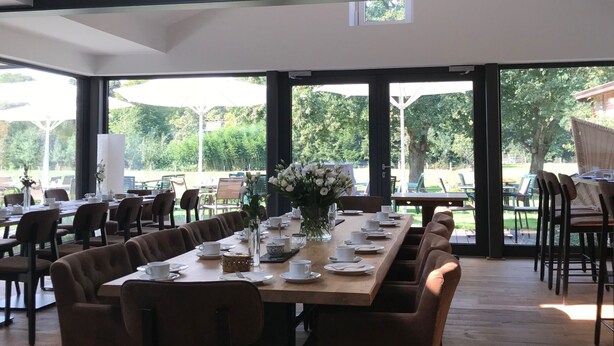 A spacious dining area with a long wooden table set for a meal, surrounded by chairs, and large windows offering a view of the outdoors. - Landcafé Neumann (Bad Zwischenahn)