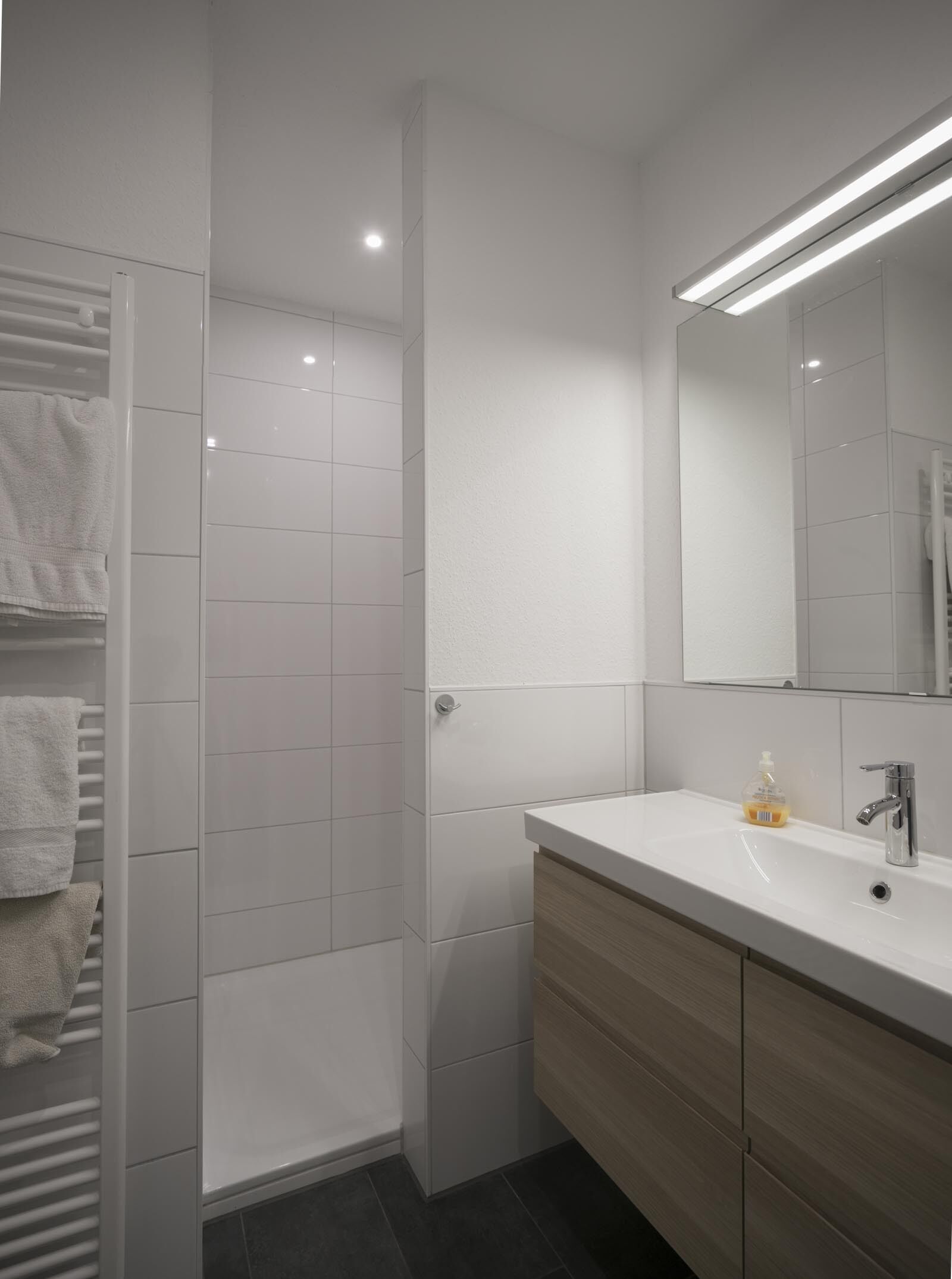 A modern bathroom with white tiles, a glass shower enclosure, a vanity with a mirror, and a towel rack.