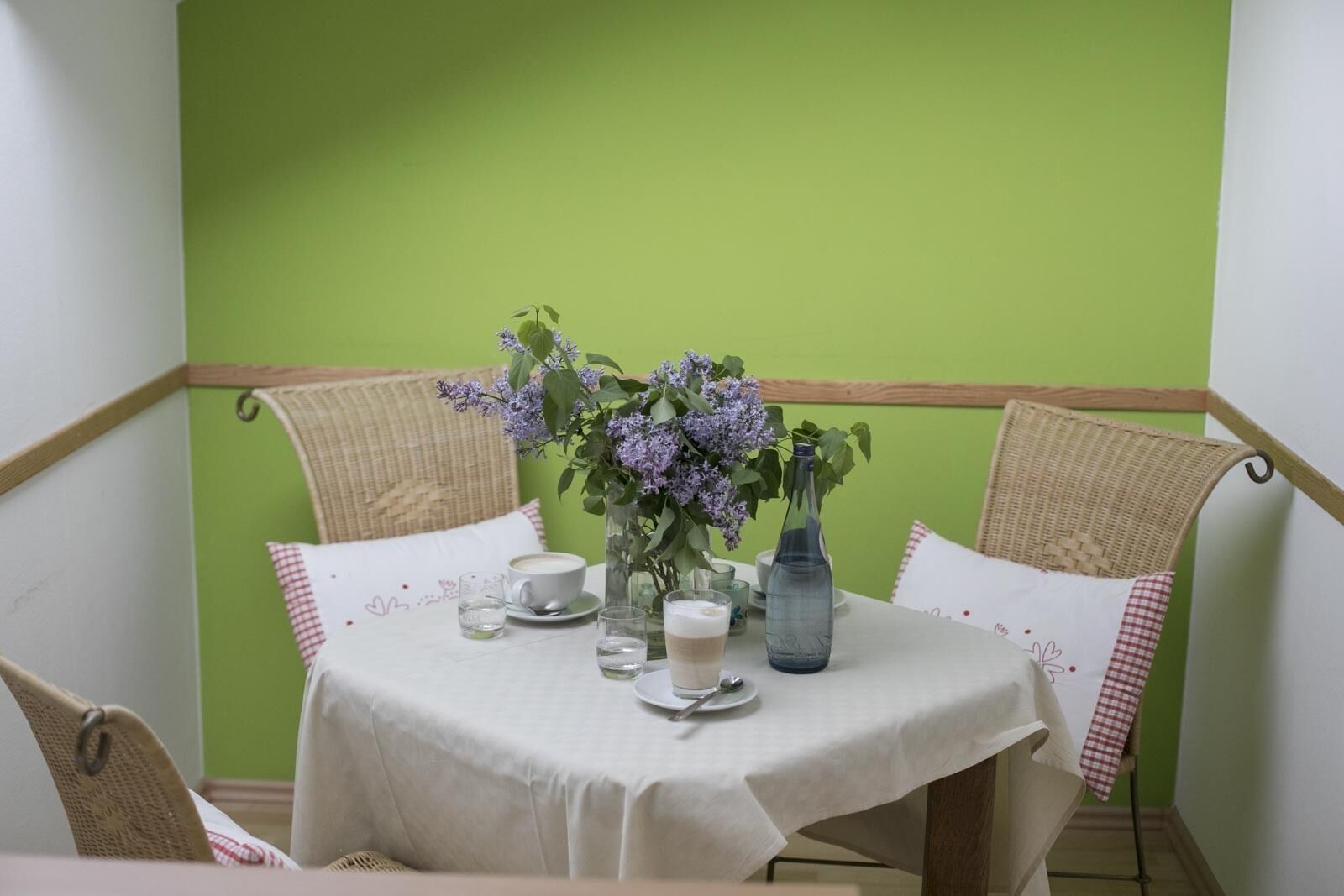 A dining table with a white tablecloth, two wicker chairs, a vase with purple flowers, a blue bottle, and a cup of coffee.