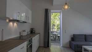 A modern kitchen with white cabinets, a wooden countertop, and a coffee maker. There is a dining area with a table and chairs, and a gray sofa. A large window with a view of trees is visible. - Landcafé Neumann (Bad Zwischenahn)
