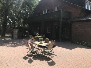 An outdoor patio with a brick floor, wooden tables, and metal chairs. - Landcafé Neumann (Bad Zwischenahn)