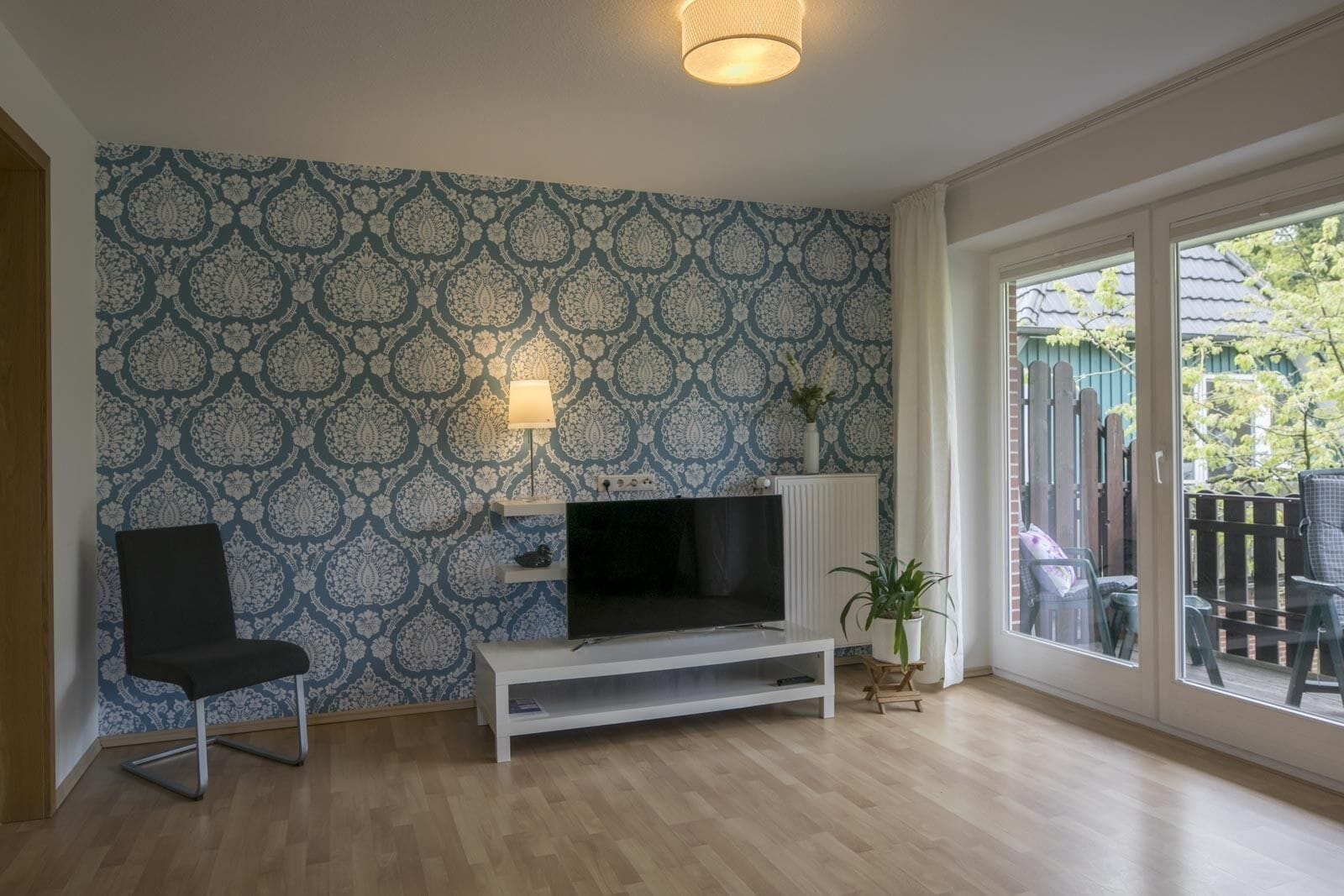 A living room with a patterned wallpaper, a TV on a white stand, a black chair, and a view of a backyard through a glass door.