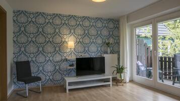 A living room with a patterned wallpaper, a TV on a white stand, a black chair, and a view of a backyard through a glass door.