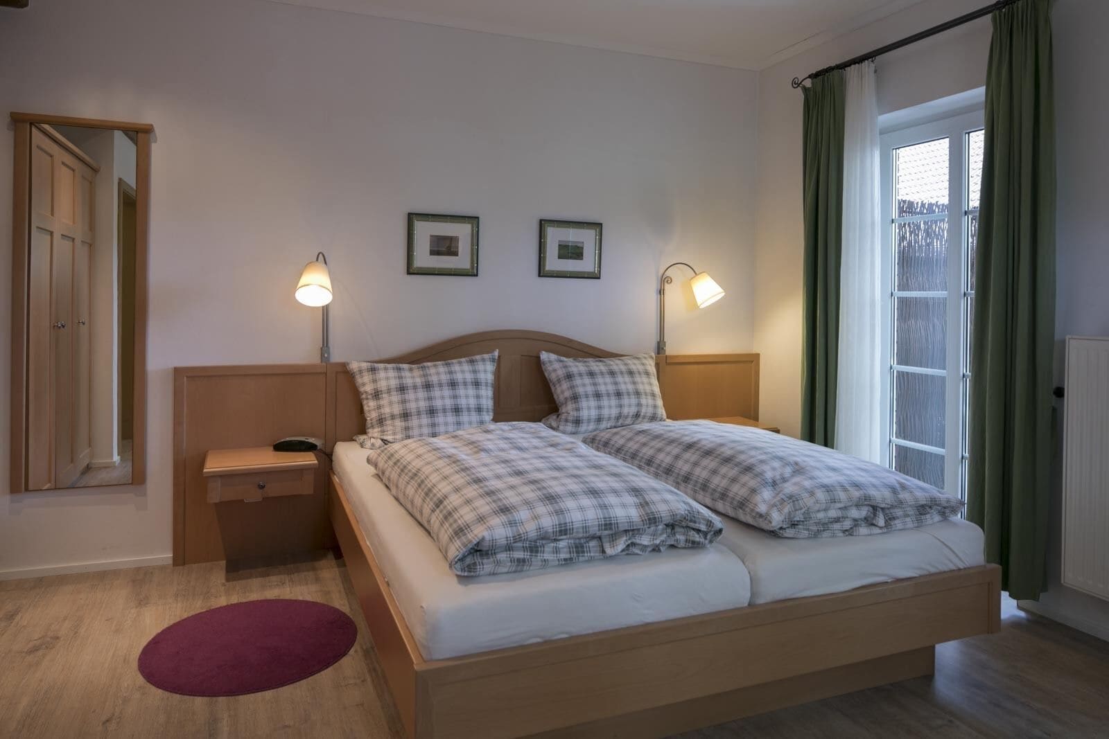 A double bed with a wooden headboard, two bedside lamps, a small bedside table, a purple rug, and a window with green curtains.
