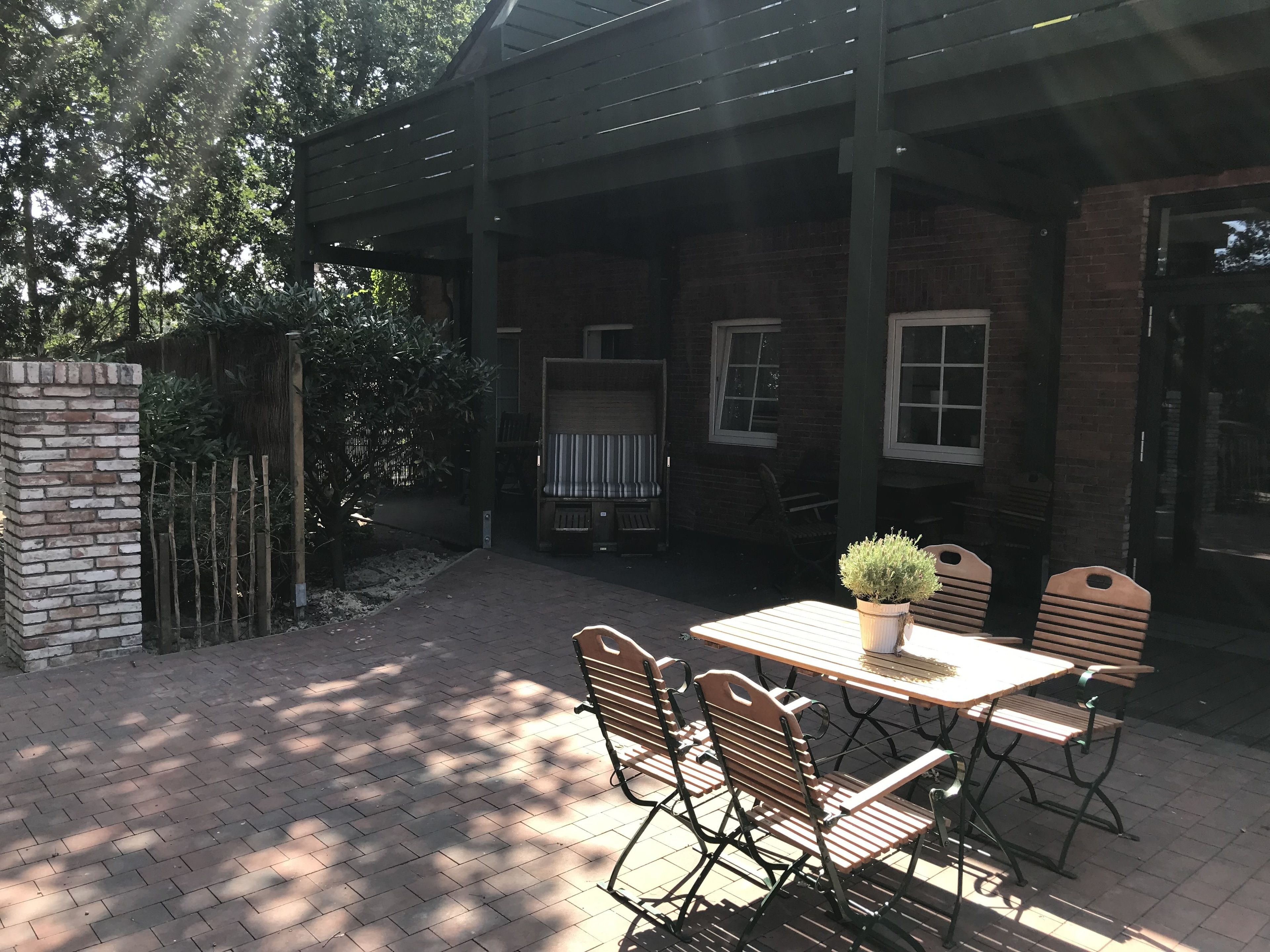 Eine Terrasse im Freien mit Backsteinfassade, Holzstühlen, einem Tisch mit Blumentopf und einem überdachten Bereich mit Fenstern und einer Bank.