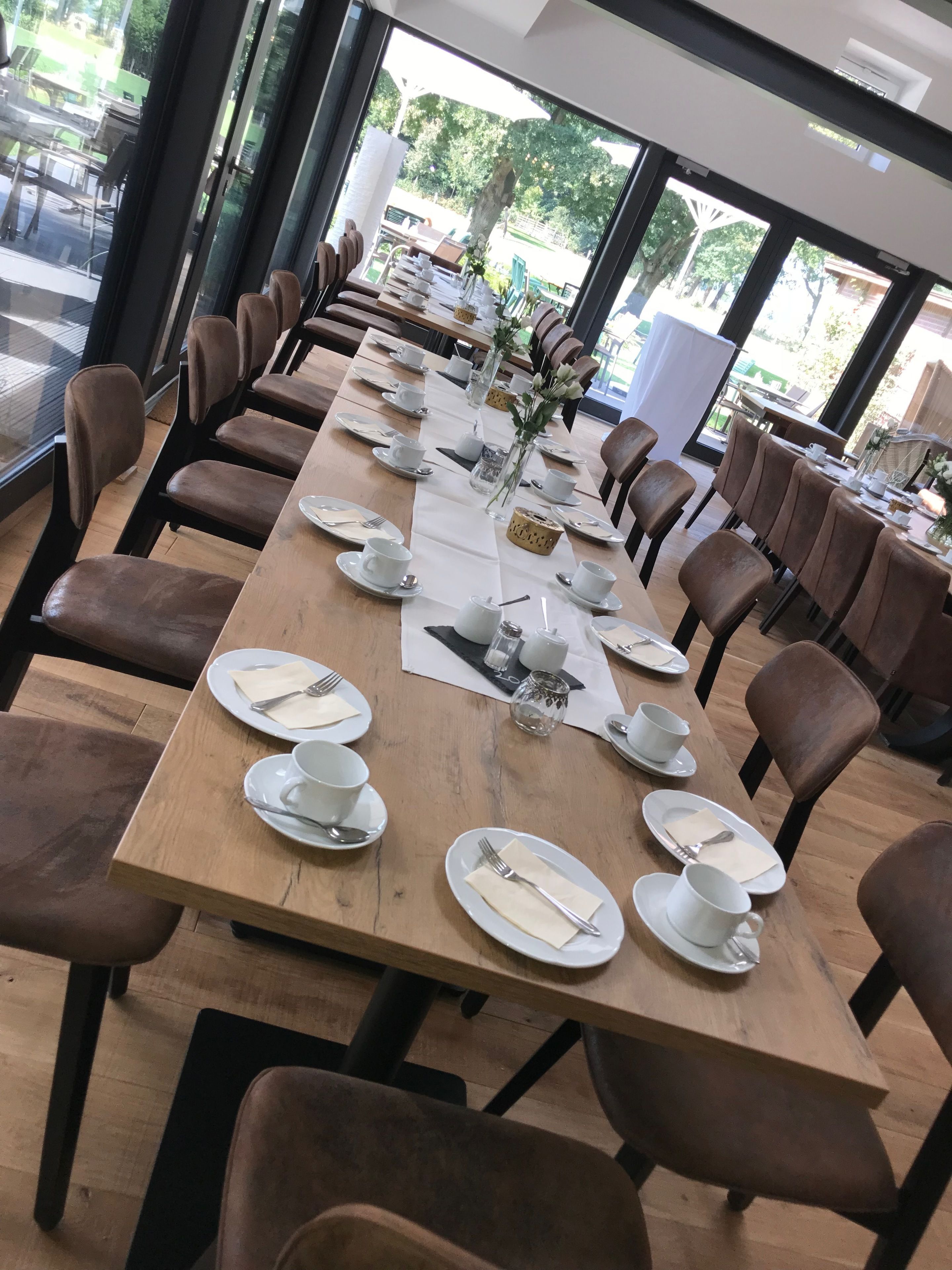 A long dining table set with white plates, cups, and cutlery, surrounded by brown chairs in a bright room with large windows.