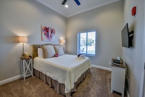 A bedroom with a large bed, two bedside lamps, a window with blinds, a flat-screen TV mounted on the wall, and a small desk with a computer.