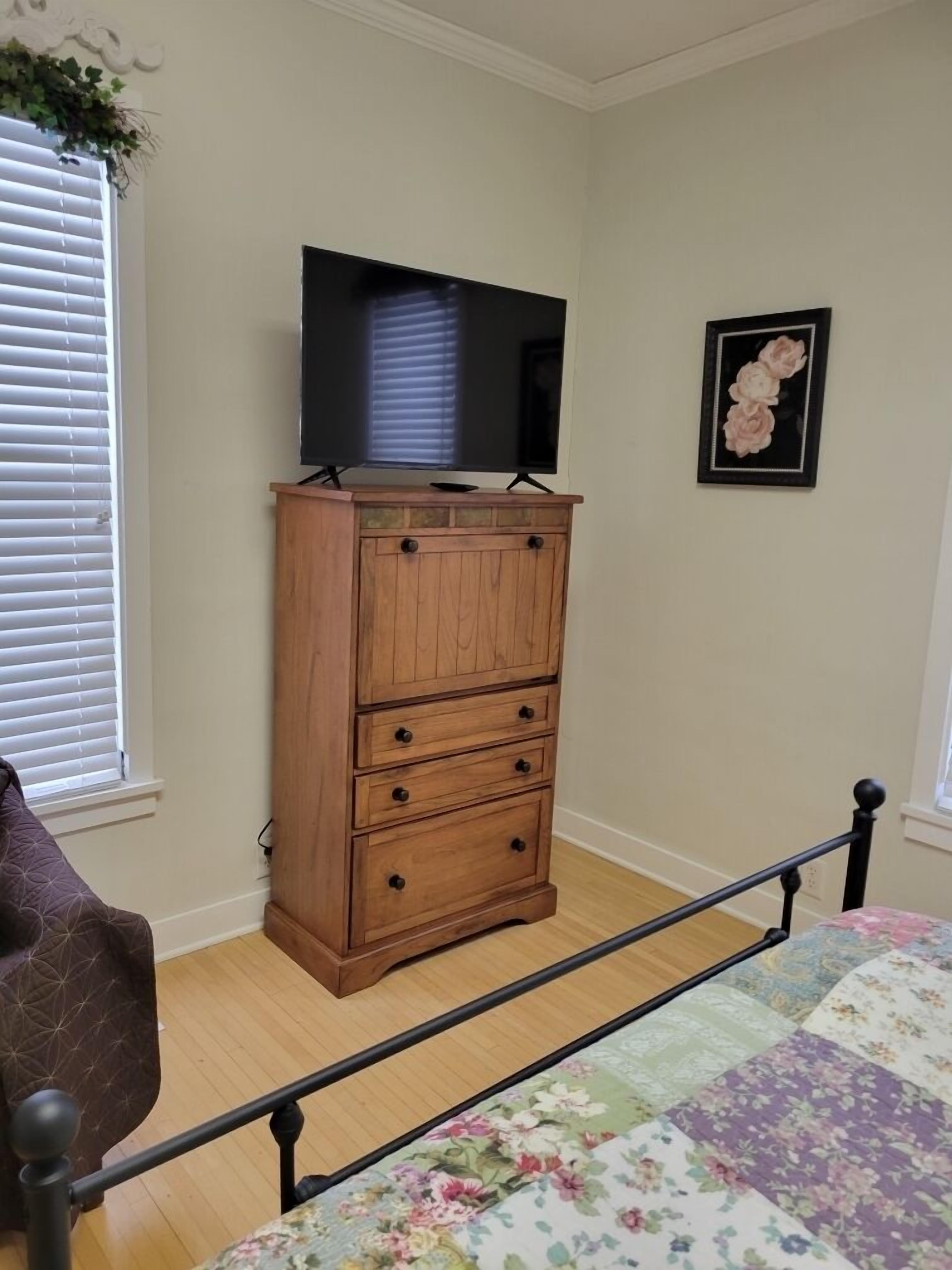 A bedroom with a wooden dresser, a bed with a floral quilt, a television on top of the dresser, and a framed floral artwork on the wall.