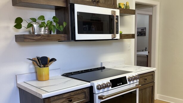 A compact kitchen with a built-in microwave, stove, and oven, wooden cabinetry, and a small shelf with kitchen utensils.
