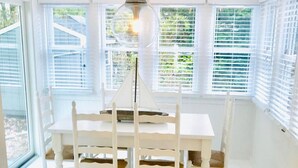 A white dining table with four chairs, a hanging light fixture, and large windows with blinds. - Renovated cottage 1 mile to beach in Chatham! (Chatham)