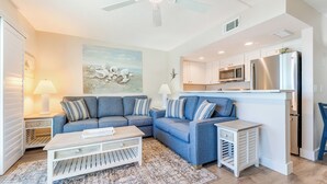 A modern living room with a blue sofa set, a white coffee table, and a kitchen area with stainless steel appliances. - OCEAN'S REACH 4B3: 3rd Floor, Gorgeous Gulf-Front Views, Amenities Open! (Sanibel Island)