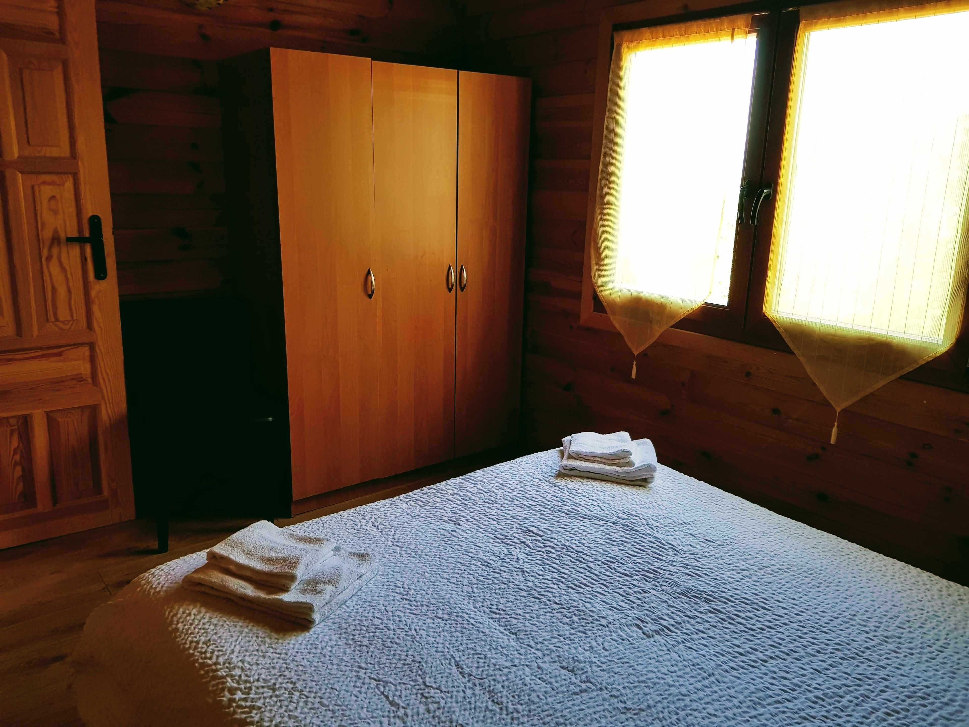 A wooden cabin room with a bed, a wardrobe, and a window with a curtain.
