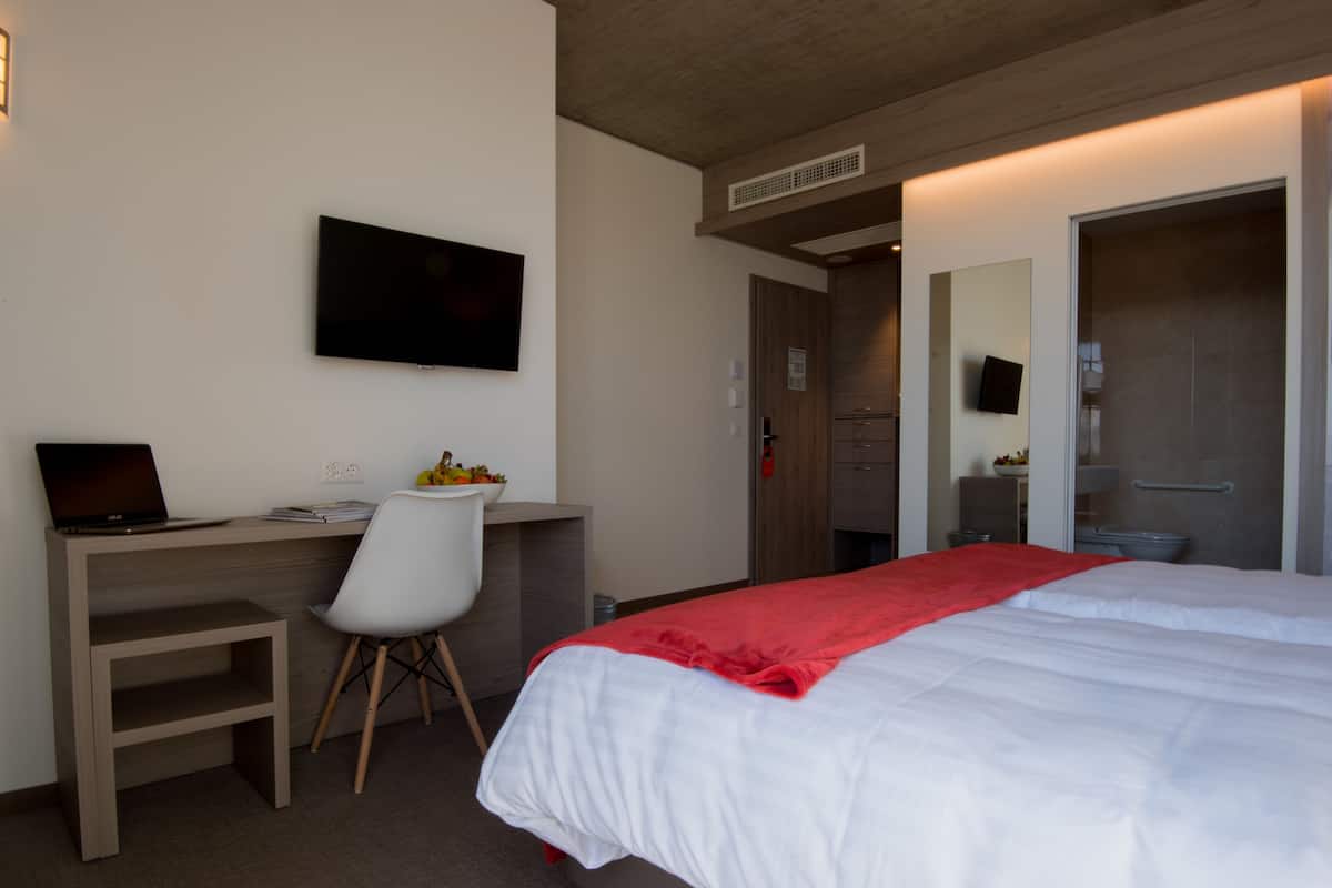 A hotel room with a bed, a desk with a laptop, a chair, and a TV on the wall.