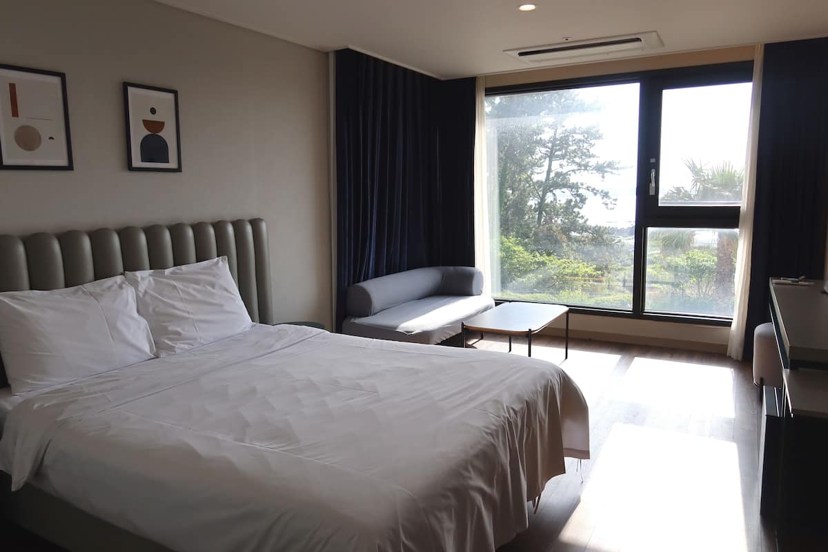 A modern hotel room with a large bed, a seating area, a desk, and a large window with a view of greenery.