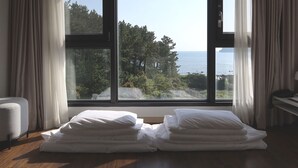 A room with a large window overlooking a forest and the sea. There are two stacks of white pillows on the floor. - Hotel Seogwipean (Seogwipo)