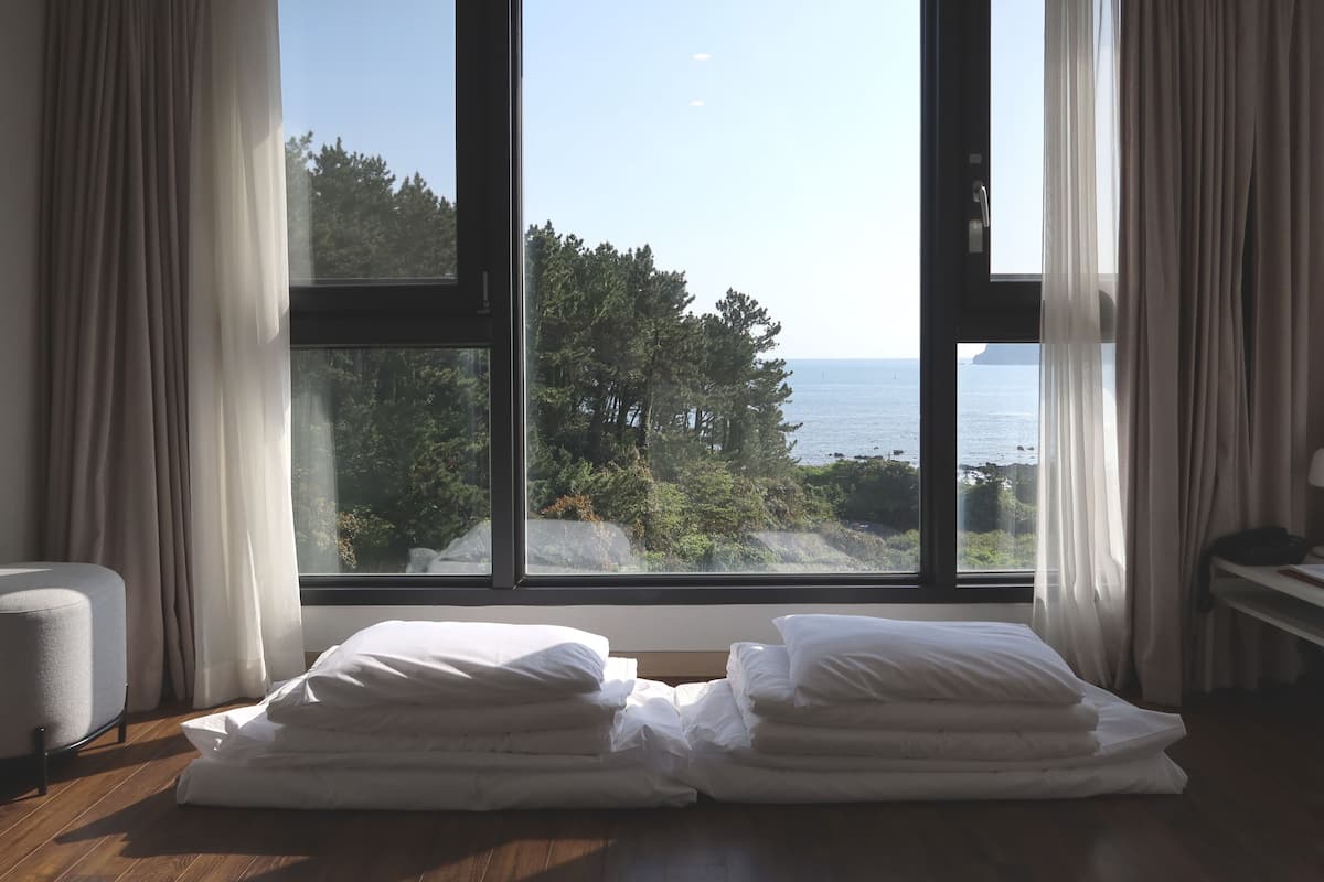A room with a large window overlooking a forest and the sea. There are two stacks of white pillows on the floor.