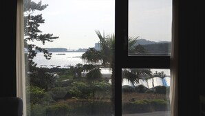 A view through a window with a view of a body of water, trees, and a distant bridge. - Hotel Seogwipean (Seogwipo)