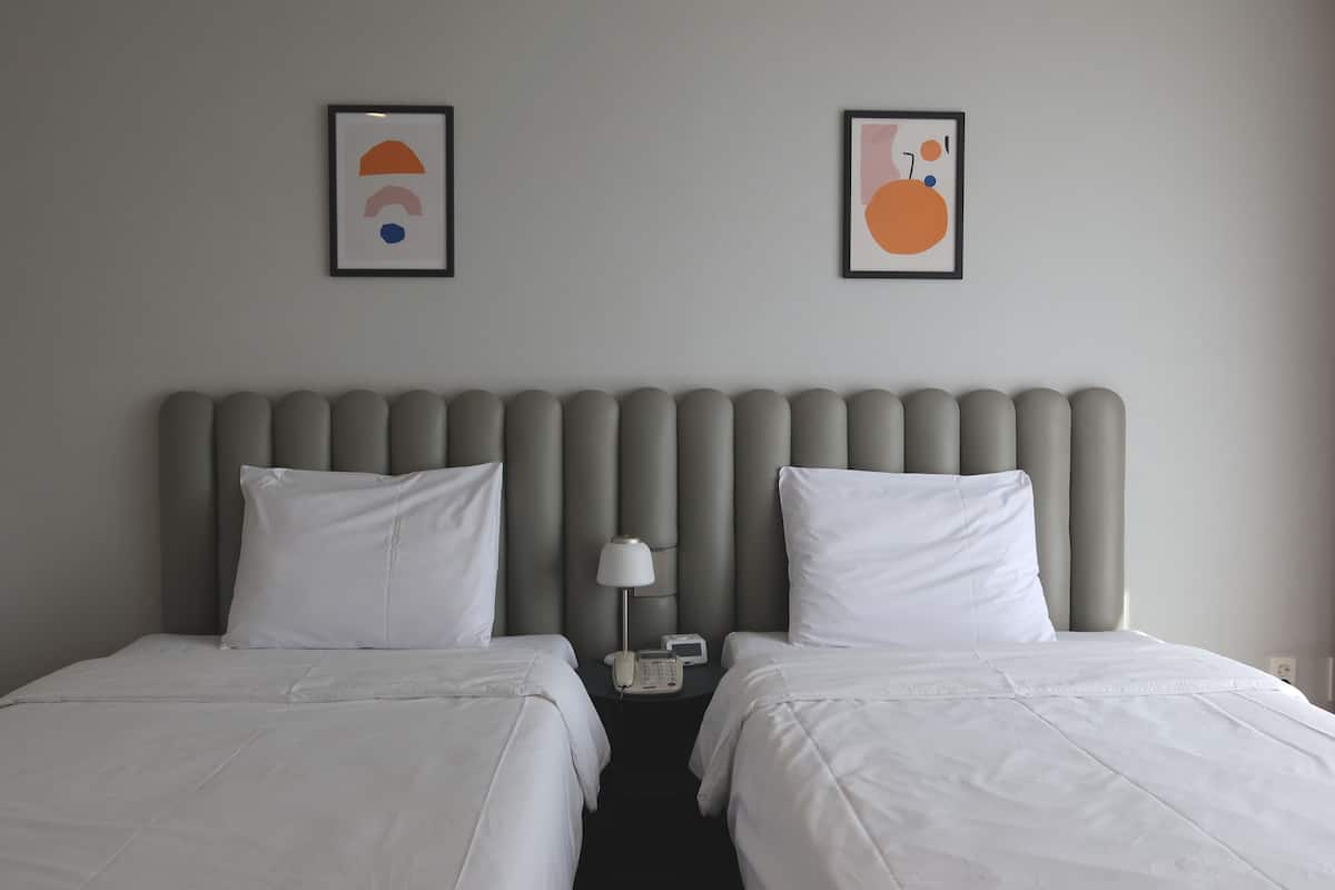 A hotel room with two beds, a grey padded headboard, white bedding, a bedside table with a lamp, and two framed artworks on the wall.