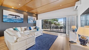 A modern living room with a beige sofa, a glass coffee table, and a blue rug. There are two paintings on the wall, a dining area with a table and chairs, and a sliding glass door leading to an outdoor space. - Saltwater 3 Apartment (Point Lookout)