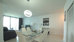 A modern dining area with a glass table, white chairs, a chandelier, and a cityscape view. - Beachfront Apt | Hollywood, 10th Floor + Pool (Hollywood)