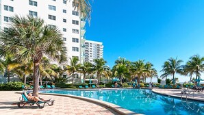 A swimming pool area with lounge chairs and palm trees, surrounded by a white building with multiple floors. - Beachfront Apt | Hollywood, 10th Floor + Pool (Hollywood)