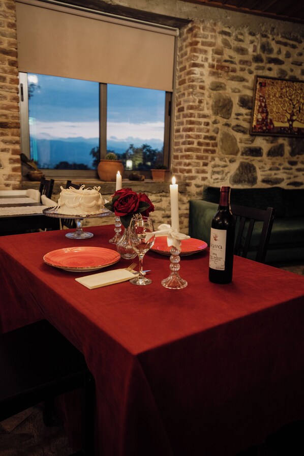 Una mesa dispuesta con pastel, vino y velas, con una pared de piedra y ventana al fondo.