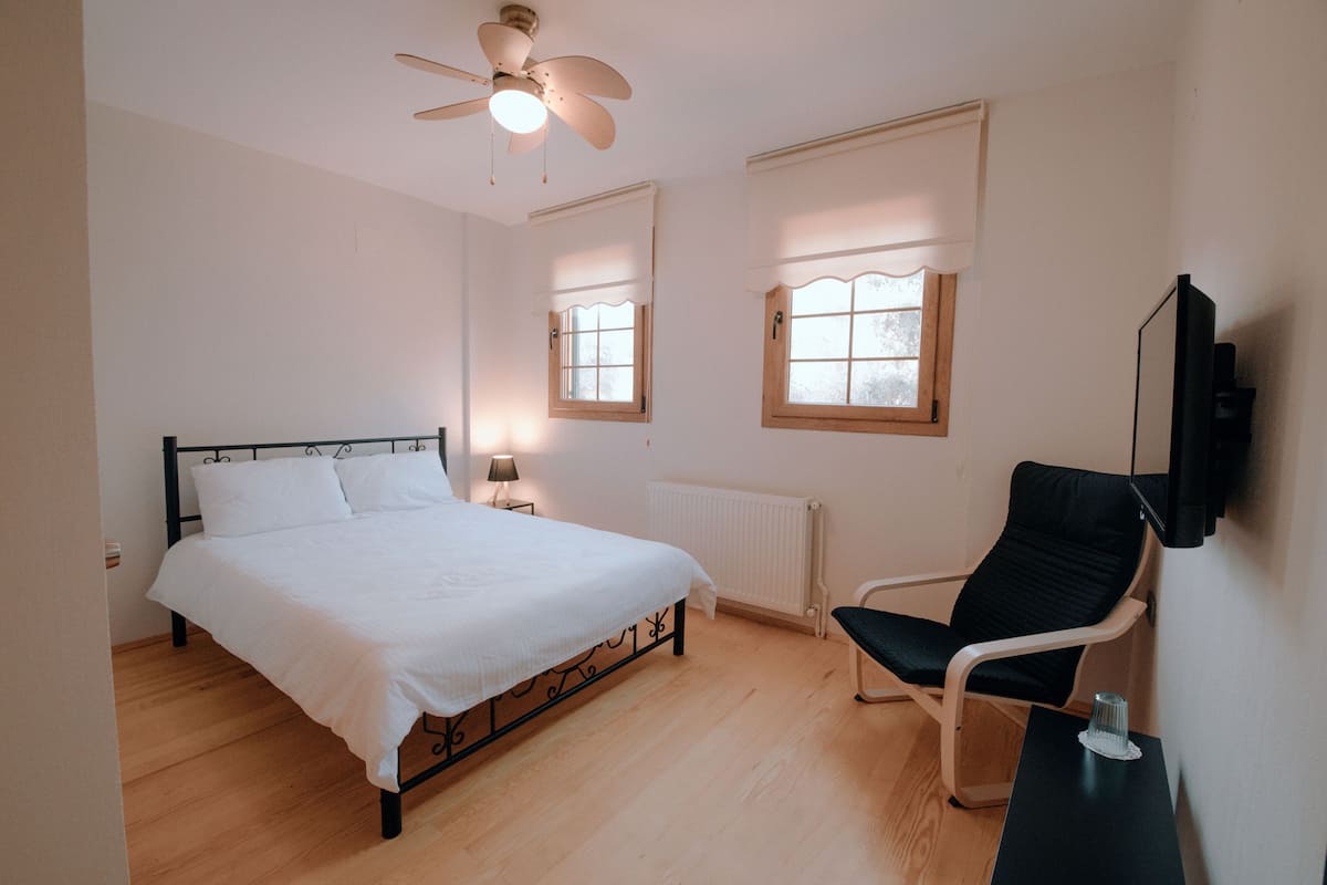 Un dormitorio con cama, una silla, ventilador de techo y dos ventanas con persianas.