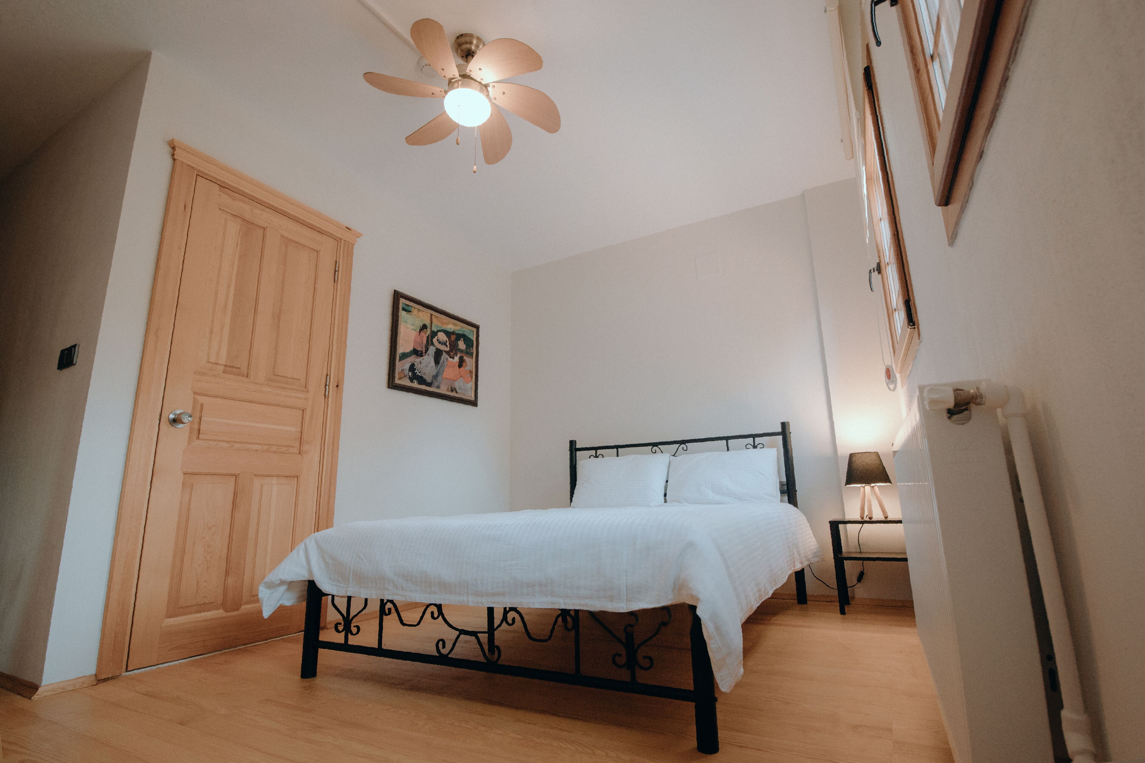 Un dormitorio con una cama, una puerta de madera, un ventilador de techo, una mesita de noche con lámpara y un cuadro en la pared.