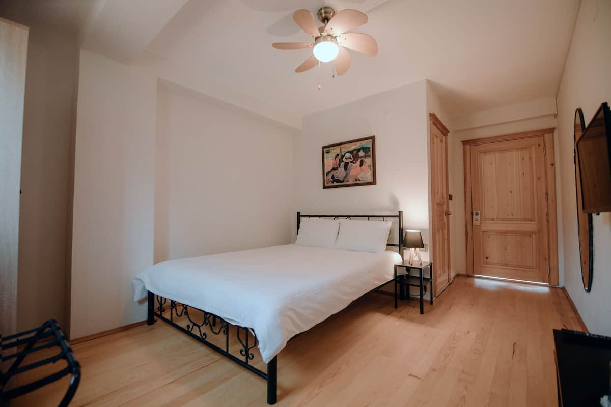 Un dormitorio con una cama, una mesita de noche, un ventilador de techo, una puerta de madera y un cuadro en la pared.