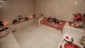 Una habitación con suelo de baldosas, una cama con una manta de estampado rojo y blanco, una mesita con velas y un jarrón con flores rojas.