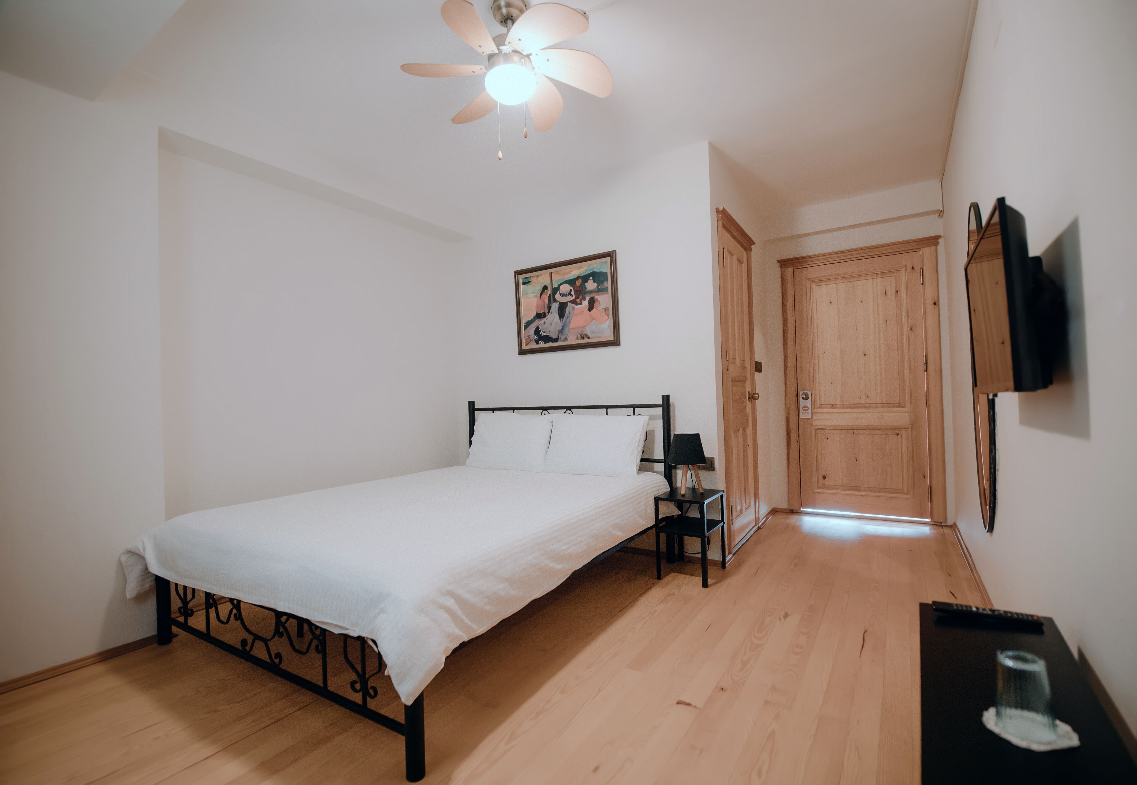 A bedroom with a bed, a nightstand, a ceiling fan, a wooden door, and a wall-mounted TV.