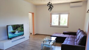 A modern living room with a flat-screen TV on a white stand, a glass coffee table, a dark brown leather sofa, and a large window providing natural light. - Ivan's holiday home (Zemunik Donji)