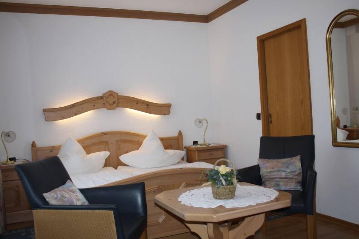 A cozy room with a wooden headboard, a bed with white pillows, a small table with a basket, and two chairs.