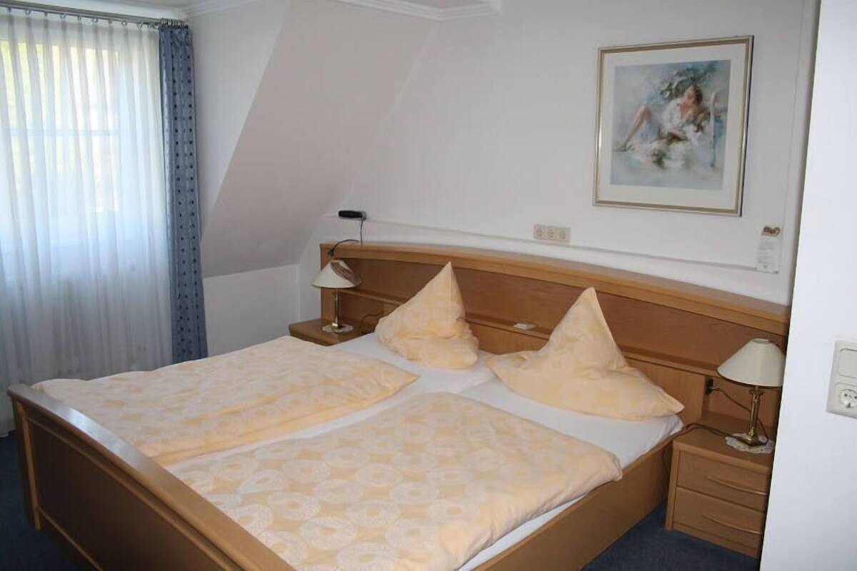 A hotel room with a double bed, two bedside tables, a nightstand, a lamp, a painting on the wall, and a window with curtains.