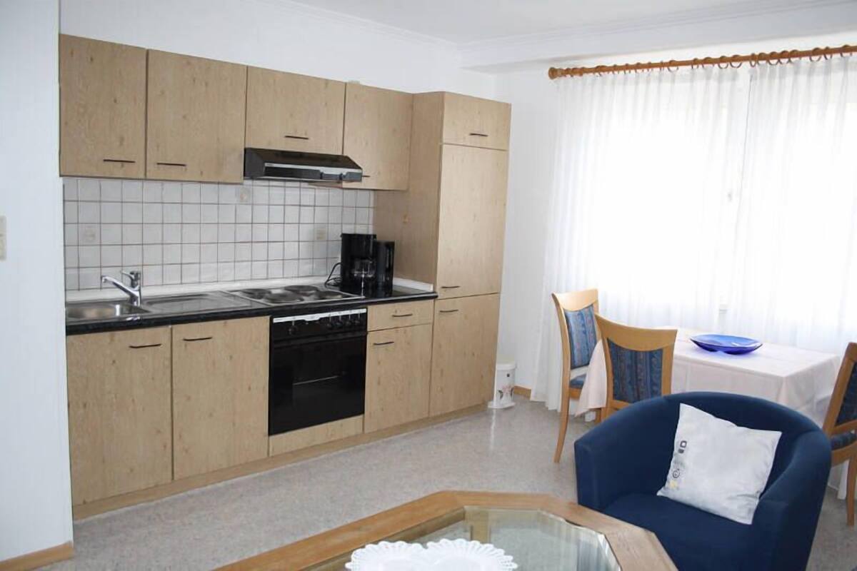 A compact kitchen with wooden cabinets, a black stove, and a coffee maker. There is a dining area with a table and chairs, and a blue chair with a white pillow.