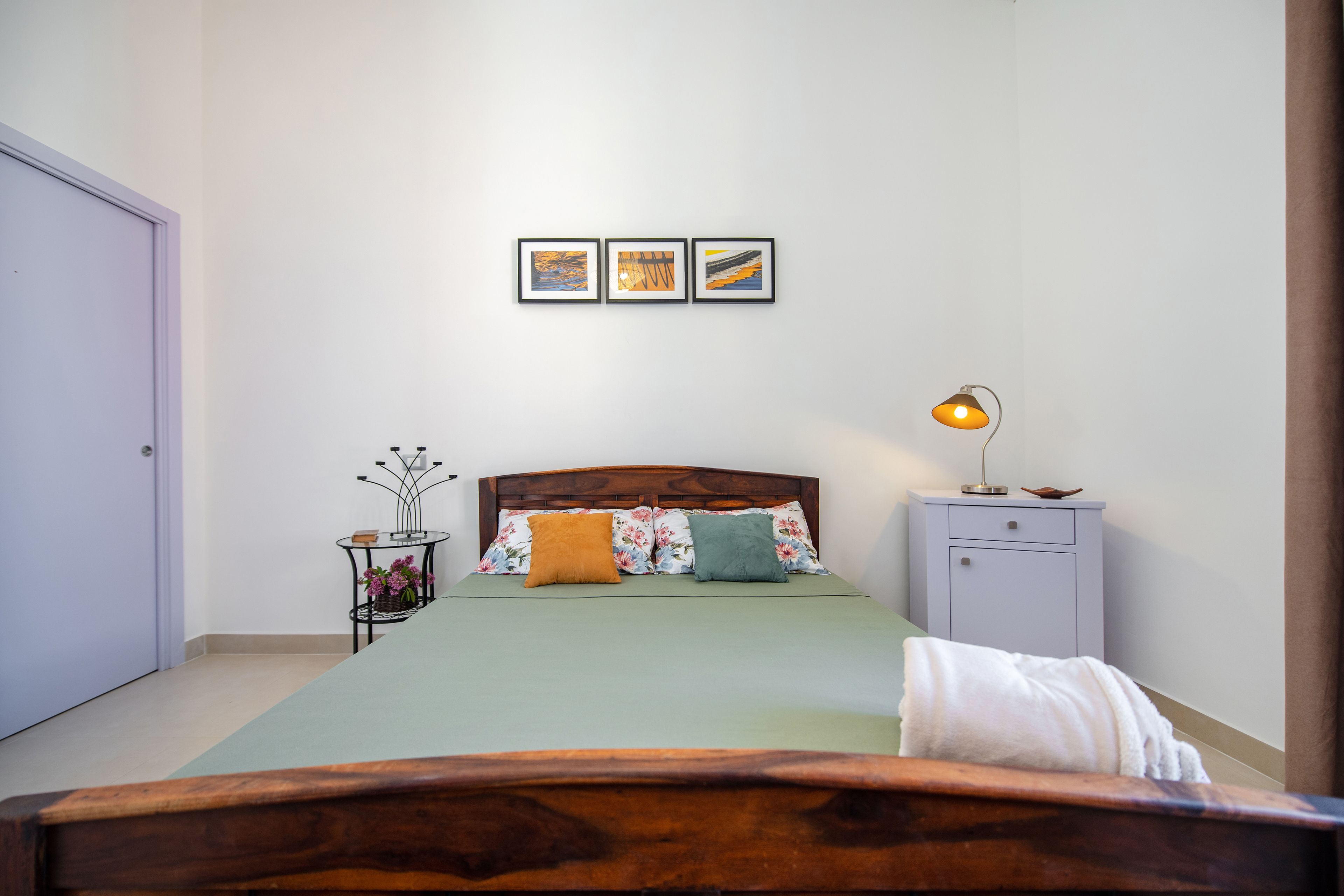 A bedroom with a wooden bed, a nightstand with a lamp, a small table with flowers, and three framed pictures on the wall.