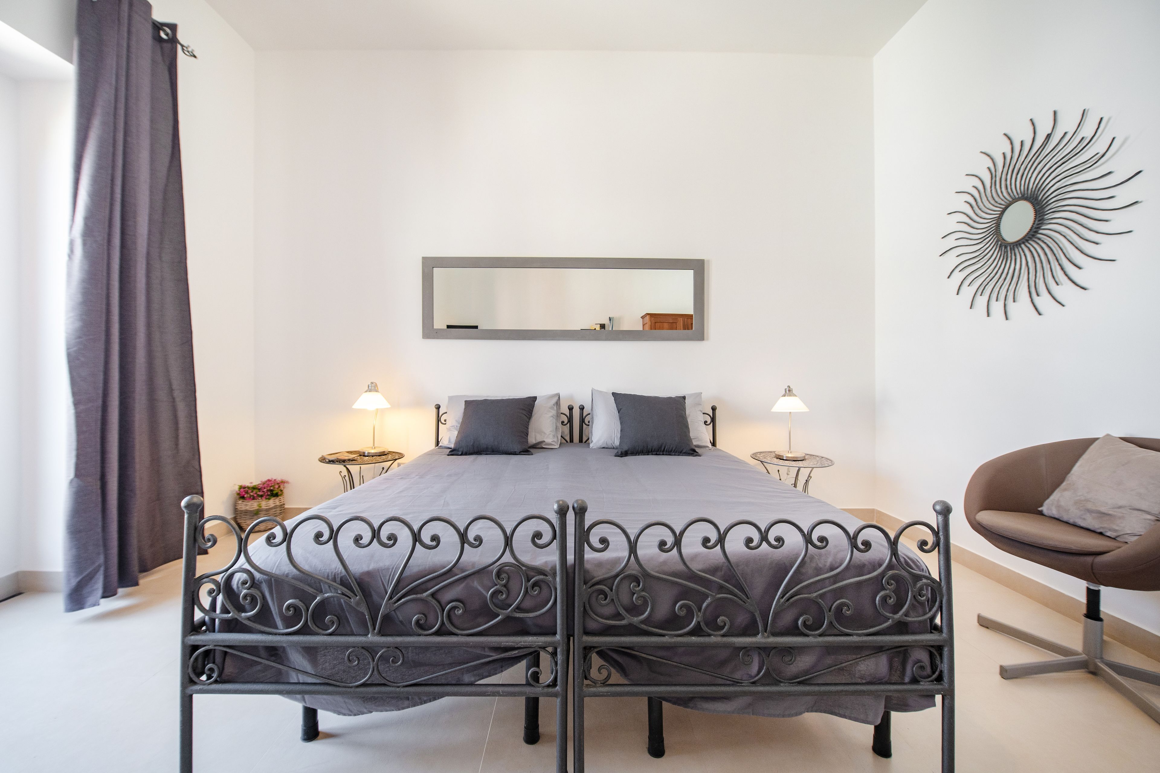 A bedroom with a bed, two bedside tables, a chair, a mirror, and a sunburst wall decoration.