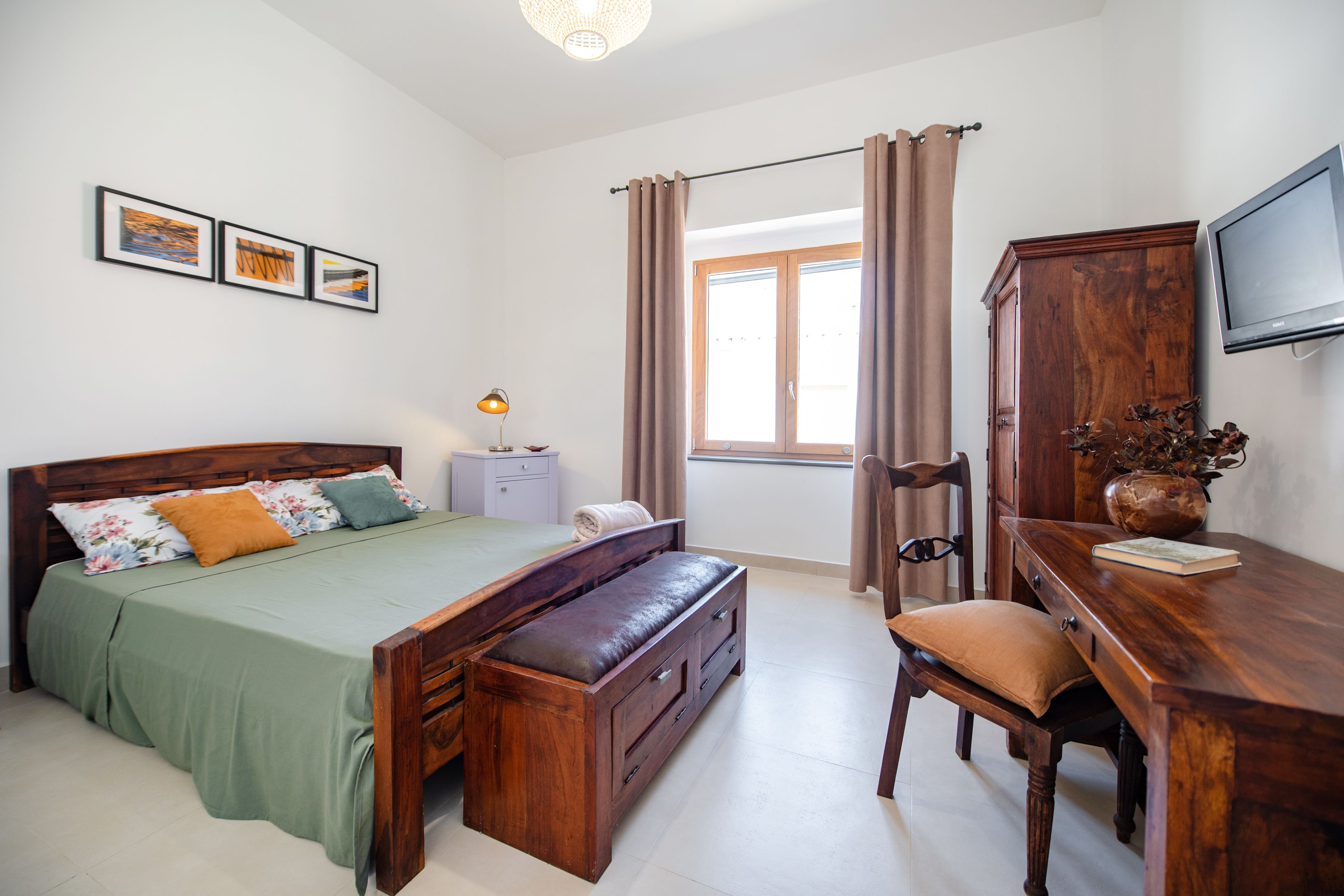 A bedroom with a wooden bed, a desk, a chair, a TV, and a window with curtains.
