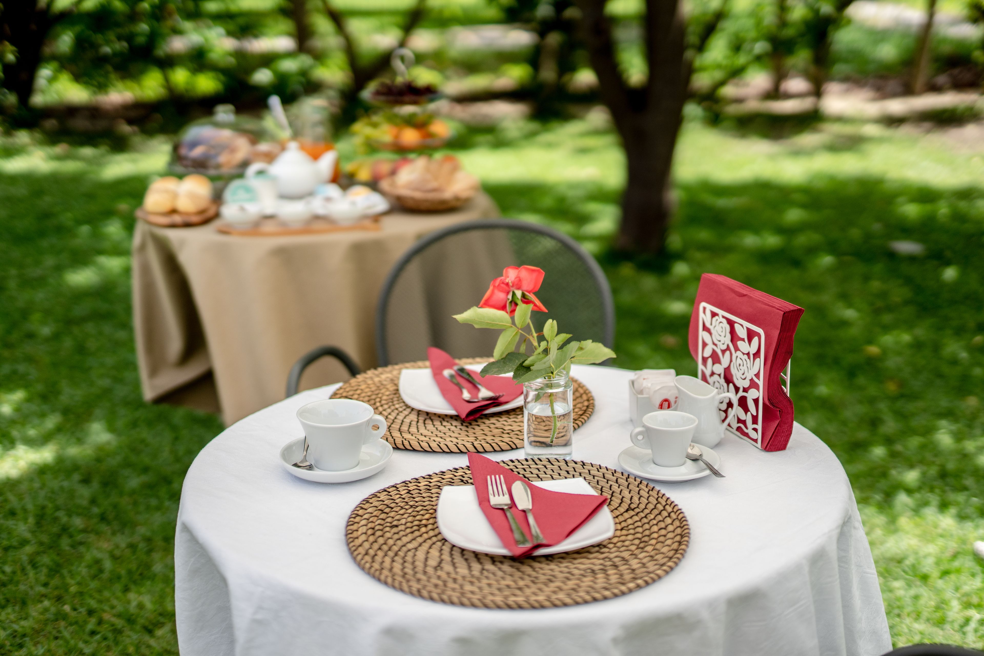 A round table set outdoors with a white tablecloth, plates, cutlery, and a red napkin.