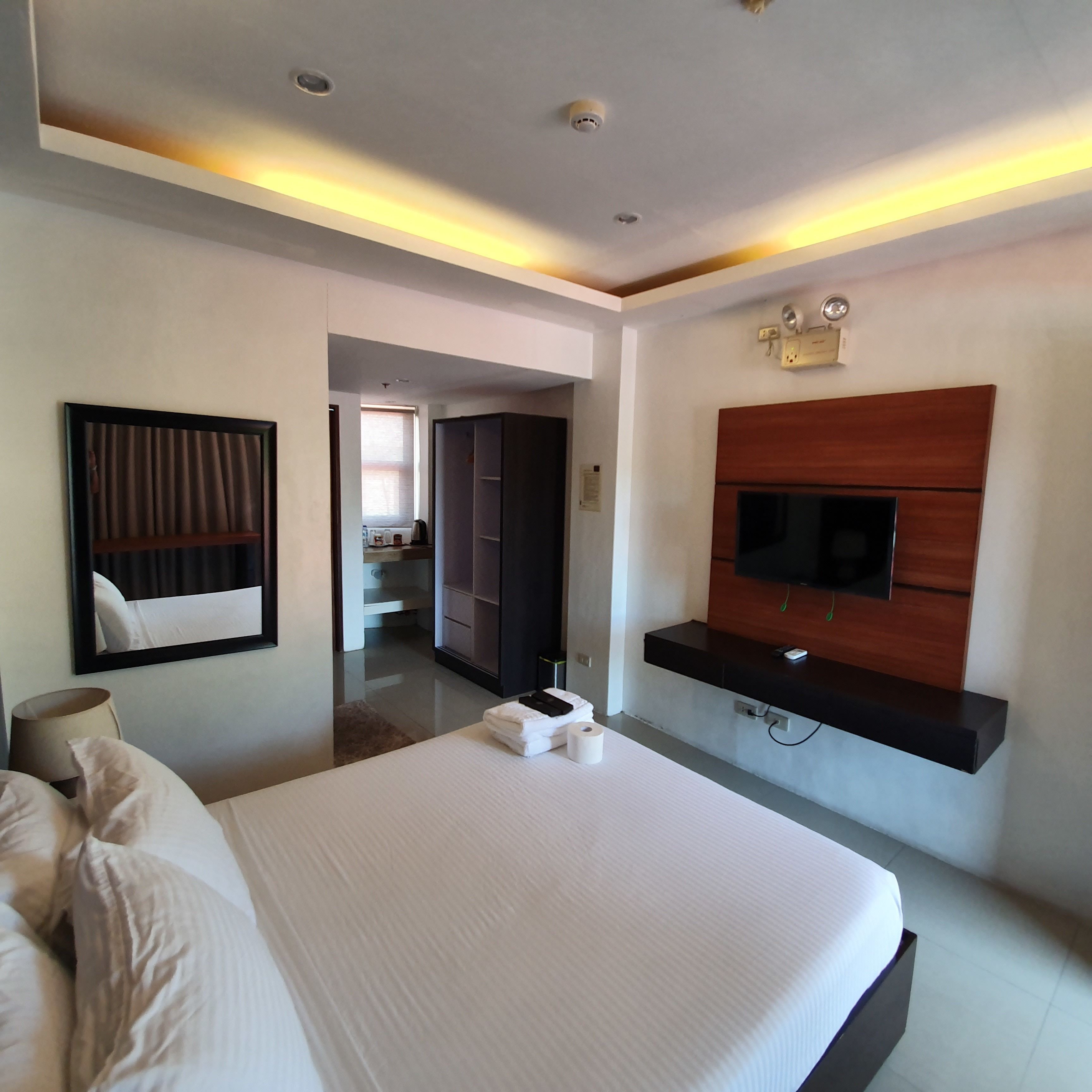 A modern hotel room with a large bed, a flat-screen TV mounted on a wooden panel, a walk-in closet, and a bathroom visible through an open door.