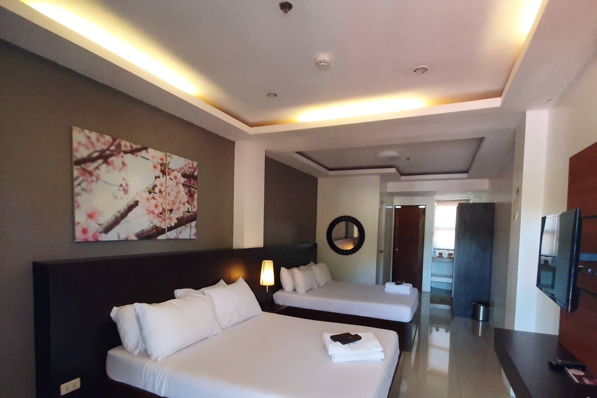 Habitación de hotel con dos camas, un cuadro grande, un televisor y un pasillo que conduce a otra habitación.