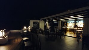 An outdoor seating area with tables and chairs, a bar area with illuminated shelves, and a statue in the background. - Hotel Zenturia (Boac)